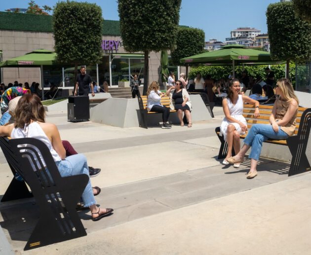 Kentsel bir plazada, siyah metal iskeletli ve doğal ahşap çıtalı modern, ergonomik STC-BANK-01 açık hava bankında oturarak sohbet eden ziyaretçiler.